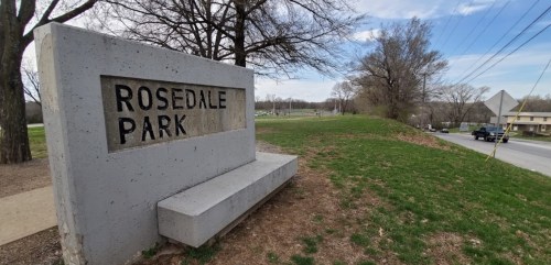 Sign for Rosedale Park with grassy area and trees in the background, alongside a road.