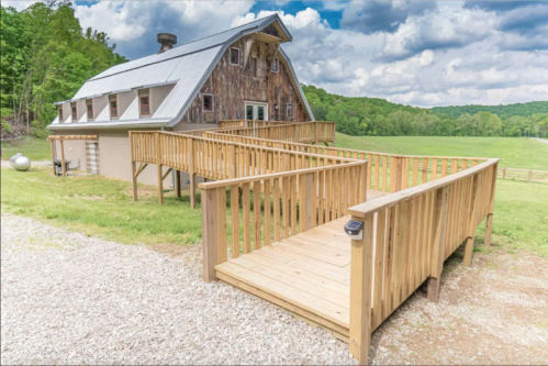 A rustic barn-style house with a wooden ramp, surrounded by green fields and a cloudy sky.