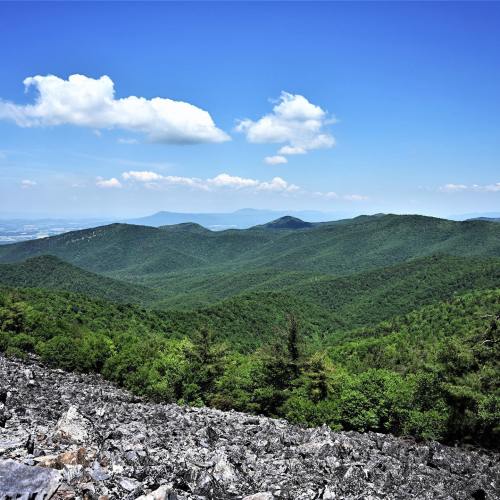 A panoramic view of lush green mountains under a clear blue sky with a few clouds. Rocky foreground adds texture.