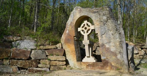 A stone arch with a carved cross stands against a backdrop of trees and rocky terrain.