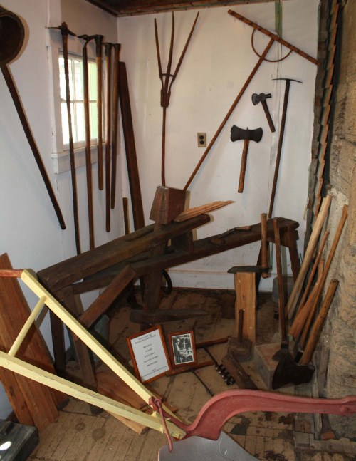 A collection of vintage farming tools and equipment displayed in a rustic room, including axes, plows, and wooden handles.