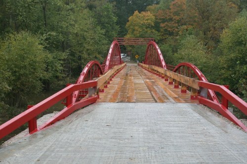 A red arch bridge spans a river, surrounded by green trees and autumn foliage. The wooden walkway is visible.