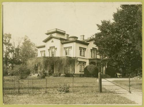 Historic two-story house with a porch, surrounded by trees and a grassy yard, featuring a decorative fence in front.