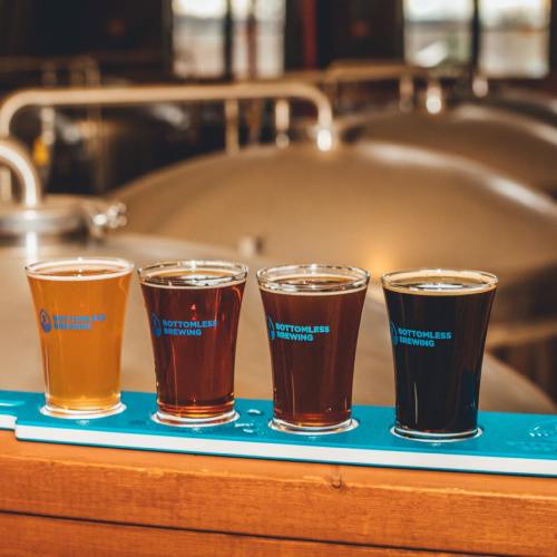 Four glasses of beer in varying shades, lined up on a wooden bar with brewing equipment in the background.