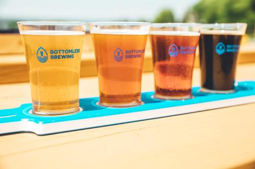 Four glasses of beer in a flight, showcasing different colors, on a wooden table with a sunny outdoor background.