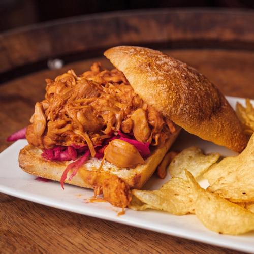 A pulled chicken sandwich on a toasted bun, served with pink slaw and crispy potato chips on the side.