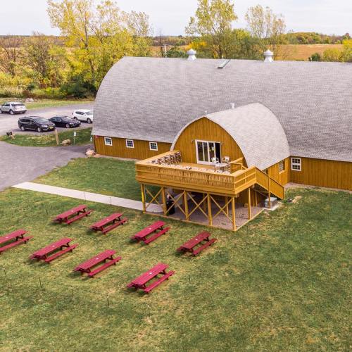 Aerial view of a rustic barn with a deck, surrounded by picnic tables and greenery, with cars parked nearby.