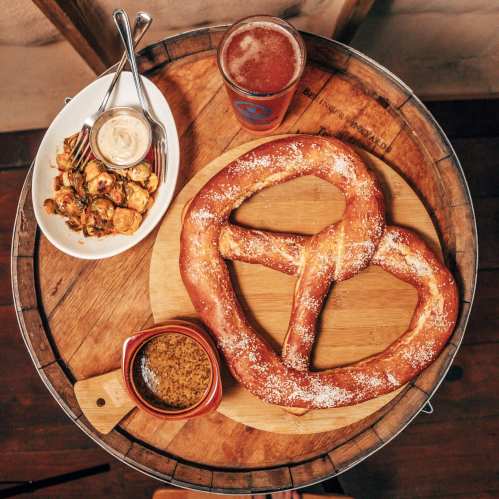 A large pretzel on a wooden board, accompanied by dipping sauces and a side of roasted vegetables, with a beer nearby.