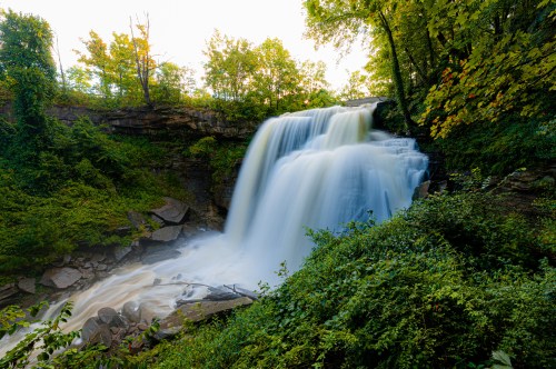A serene waterfall cascades over rocks, surrounded by lush green trees and foliage in a tranquil natural setting.