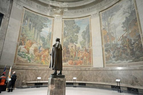 A statue stands in a grand hall, surrounded by murals depicting historical scenes on the walls.