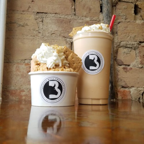 Two dessert cups on a wooden table: one with ice cream topped with whipped cream and a cookie, the other a milkshake.