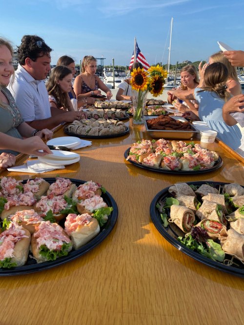 A group of people enjoying a buffet of sandwiches and wraps on a sunny day by the water, with sunflowers in the center.