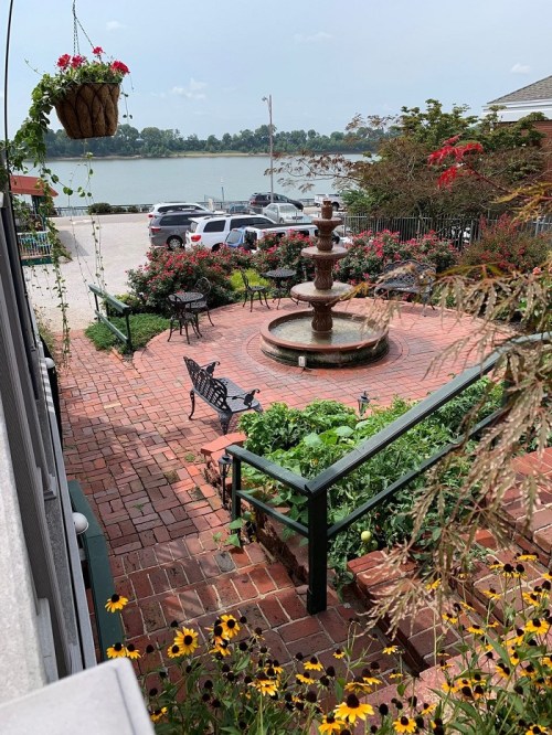 A brick patio with a fountain, surrounded by flowers and greenery, overlooking a calm body of water and parked cars.