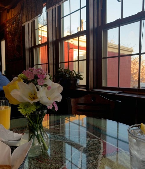A table with a vase of flowers, a glass of water, and a view of colorful buildings through large windows.