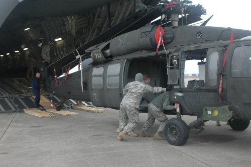 Military personnel maneuver a helicopter onto a transport aircraft on a loading ramp.