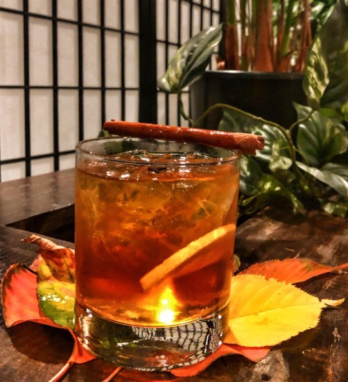 A glass of amber-colored cocktail garnished with a cinnamon stick, resting on colorful autumn leaves.