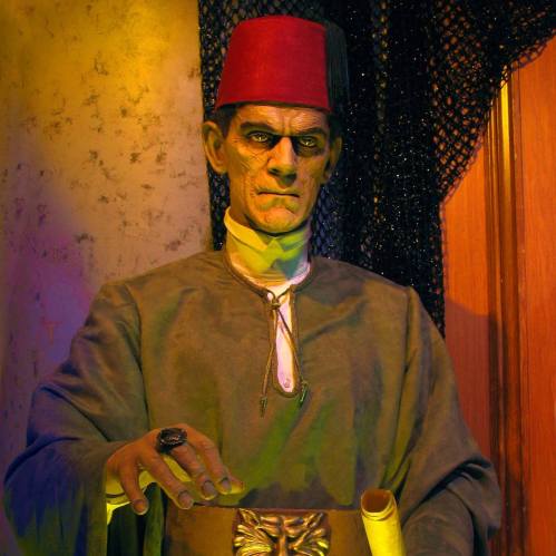 A creepy figure in a fez, wearing a gray robe, holding a scroll, with an eerie expression in a dimly lit setting.