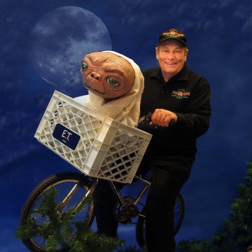 A person smiles while riding a bike with a life-sized E.T. figure in a basket, set against a moonlit backdrop.