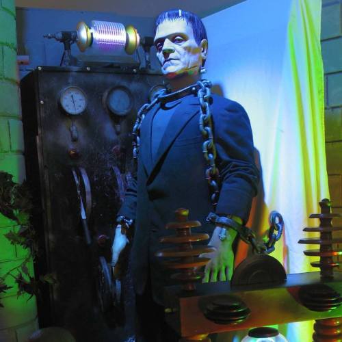 A life-sized figure resembling a classic monster, chained and standing next to vintage lab equipment with colorful lighting.