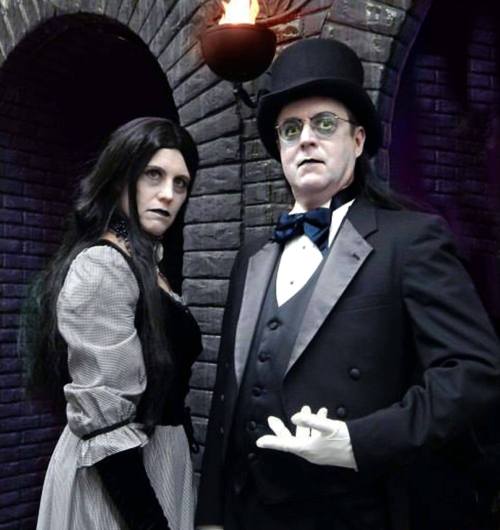 A man in a tuxedo and top hat stands beside a woman in a Victorian dress, both posing dramatically in a dimly lit archway.