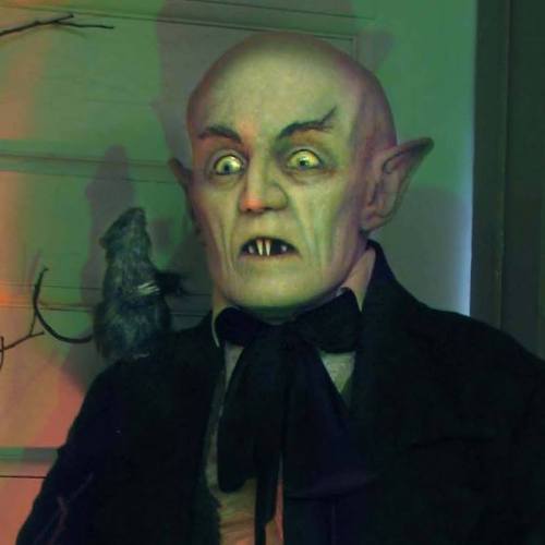 A creepy figure with pointed ears and sharp teeth stands against a wall, with a rat perched on its shoulder.