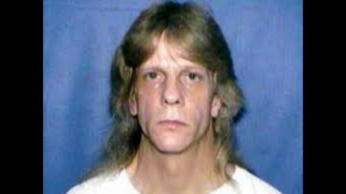 A man with long hair and a serious expression poses for a mugshot against a blue background.