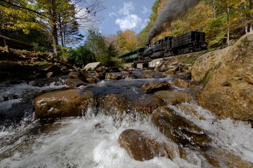A steam train travels alongside a rocky stream, surrounded by trees and autumn foliage under a blue sky.