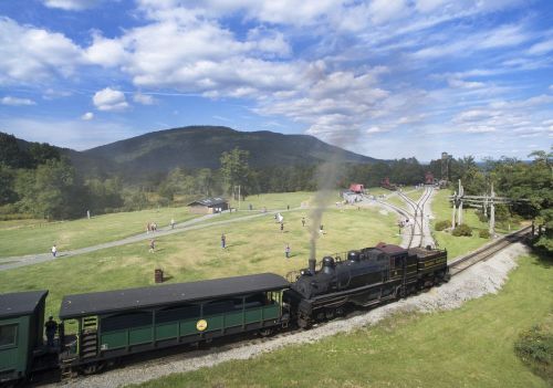 A steam train travels along tracks with people enjoying a grassy area and mountains in the background under a blue sky.