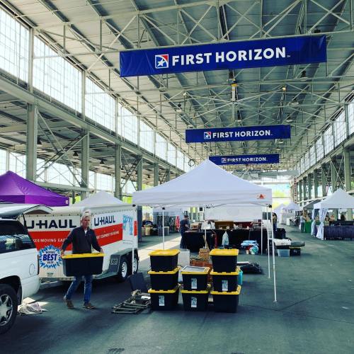 A vendor carries a box at a market with tents and banners for First Horizon and U-Haul in a spacious indoor venue.