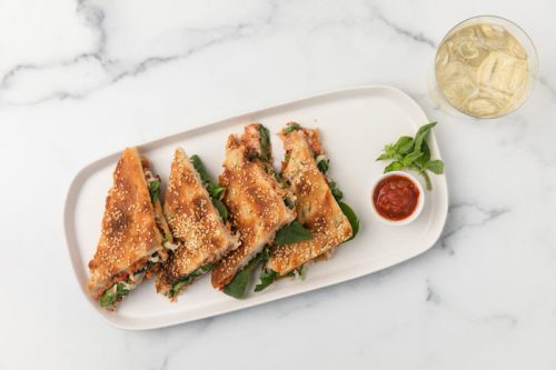 A plate of crispy, sesame-topped sandwiches filled with greens, served with a small bowl of sauce and a drink.