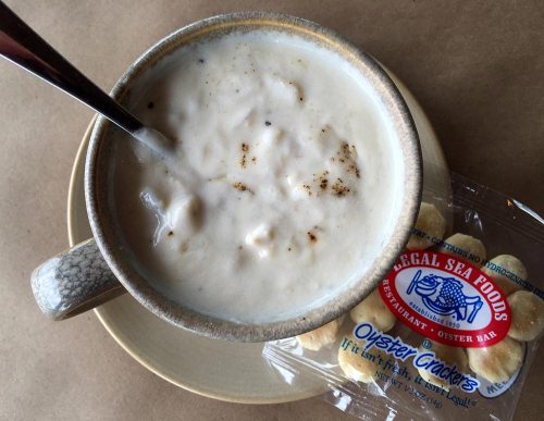 A bowl of creamy clam chowder with a spoon, accompanied by a pack of oyster crackers on a plate.