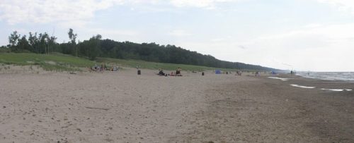 A sandy beach with people relaxing, trees in the background, and a calm lake under a cloudy sky.