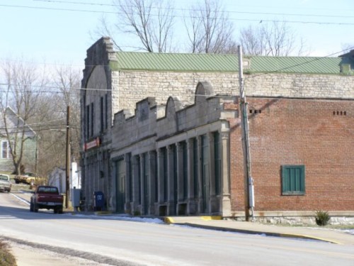 Historic stone and brick buildings line a quiet street, with a green roof and a few parked cars nearby.