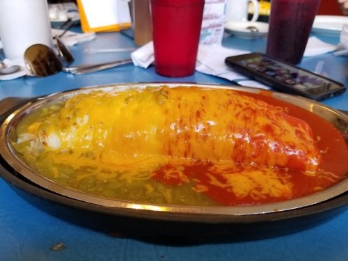 A large, cheesy enchilada topped with red and green sauces, served on a sizzling plate in a restaurant setting.