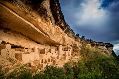 Ancient cliff dwellings nestled under a rocky overhang, surrounded by trees and a dramatic sky.