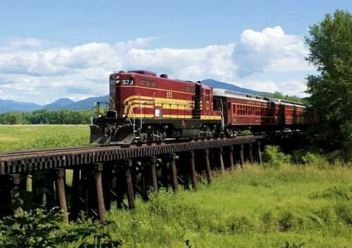 A vintage red and yellow train travels across a wooden trestle bridge, surrounded by green fields and mountains.