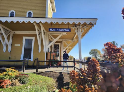 A person stands outside the North Conway train station, featuring yellow architecture and colorful flowers in the foreground.