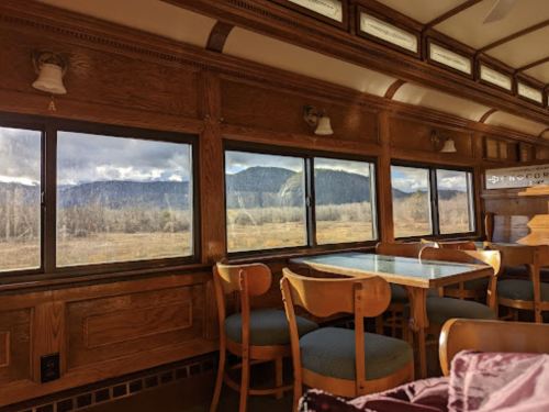 Interior of a vintage train car with wooden paneling, a table, and large windows showcasing a mountain view.