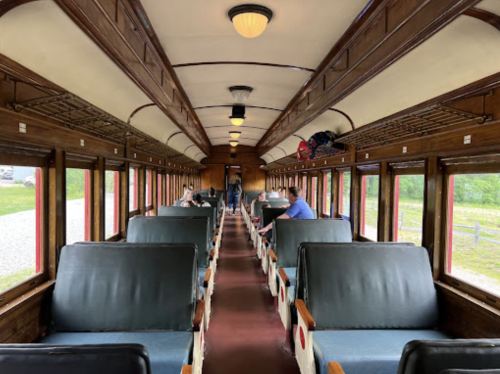 Interior of a vintage train car with wooden paneling, green seats, and passengers seated along the aisle.