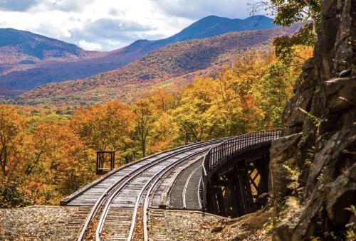 A winding railway track curves through vibrant autumn foliage and mountains under a partly cloudy sky.