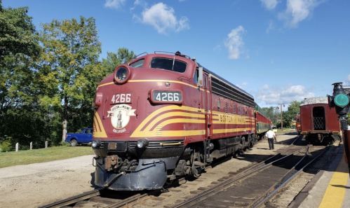 A vintage red and yellow train at a station, with a person walking nearby and trees in the background.