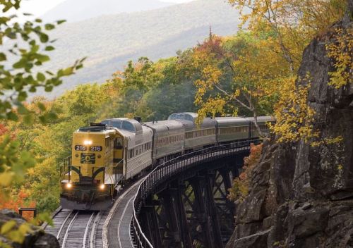 A yellow and silver train travels along a curved bridge surrounded by vibrant autumn foliage and mountains.