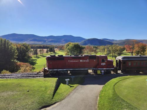 A red train passes through a scenic landscape with mountains and colorful autumn trees under a clear blue sky.