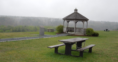 A gazebo in a misty landscape with a picnic table in the foreground and greenery in the background.