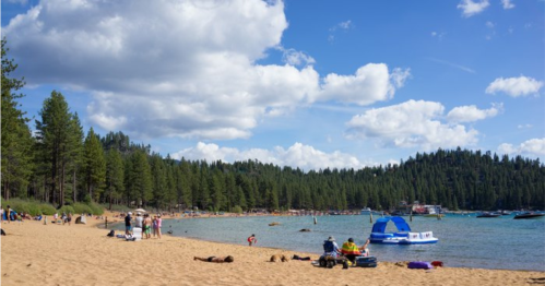 A sandy beach by a lake, with people enjoying the sun, trees in the background, and boats on the water.