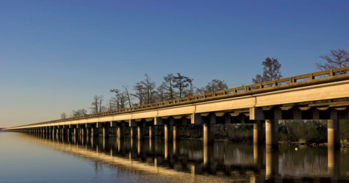 A long bridge stretches over calm water, reflecting the clear blue sky and surrounding trees.