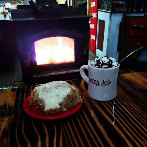 A warm cinnamon roll with icing on a red plate next to a mug of hot chocolate topped with whipped cream, by a cozy fire.