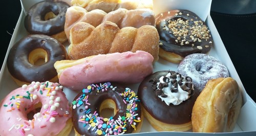 A box of assorted donuts, including glazed, chocolate, and frosted varieties, topped with sprinkles and nuts.