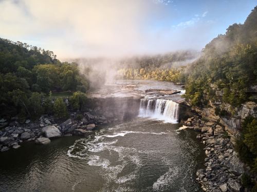 A serene waterfall cascades into a river, surrounded by lush greenery and misty morning fog.