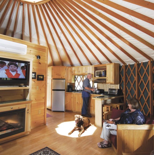 A cozy yurt interior with a man cooking, a woman sitting with a dog, and a TV in the background. Warm wooden decor.